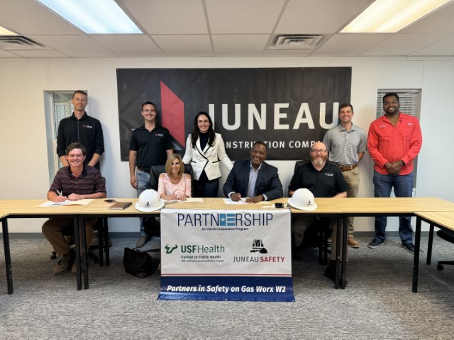 The 4 signatories seated at a table sign the agreement while 5 attendees standing behind them observe the signing. 