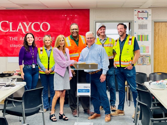 Tampa Area Office Director, OSHA: and Clayco Construction CEO stand behind a podium, with Clayco Regional Safety Managers, and USF On-site Consultation Program Director standing behind them, after signing the agreement.  