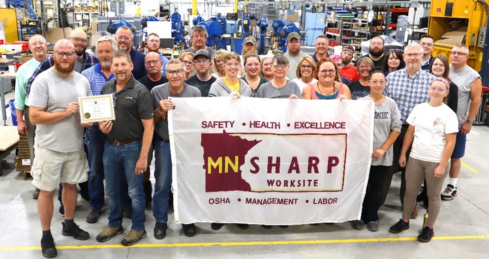 Gemini Inc. employees stand on their shop floor, proudly holding their SHARP flag