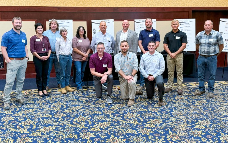 In a hotel conference room, 3 men are kneeling with 10 people standing behind them. They are, OSHA staff, Partnership signatories, supporting company and labor representatives, assembled for a smaller group photo after the signing ceremony.