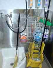 mop and bucket next to a floor sink with a broom inside. Taps with hoses are connected to a cleaning chemical dispenser.