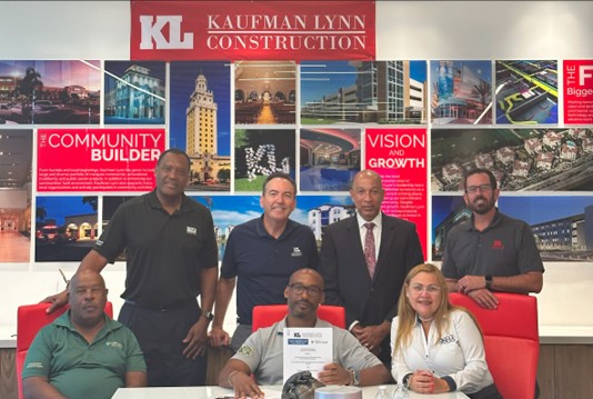Three people are seated at a table and four people stand behind them. They are representatives from OSHA’s Ft. Lauderdale Area Office, Kaufman Lynn Construction and USF SafetyFlorida Consultation, after signing the agreement.