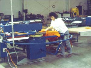 Figure 16. Automated screen printing operation. Woman leaning forward to pull and stretch a t-shirt for printing.