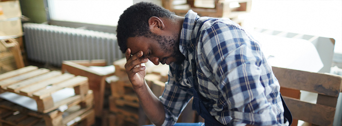 A middle-aged worker in a checked shirt sits in a room full of pallets and tools, holding his head in his hand with a stressed expression on his face.